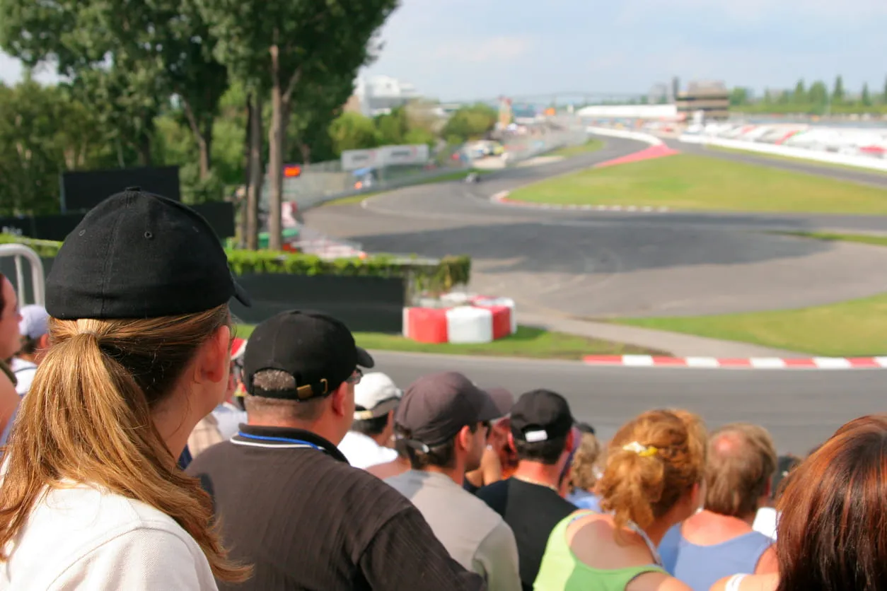 Crowd of spectators watching a race on a sunny day at a motorsport track with a curving asphalt course.