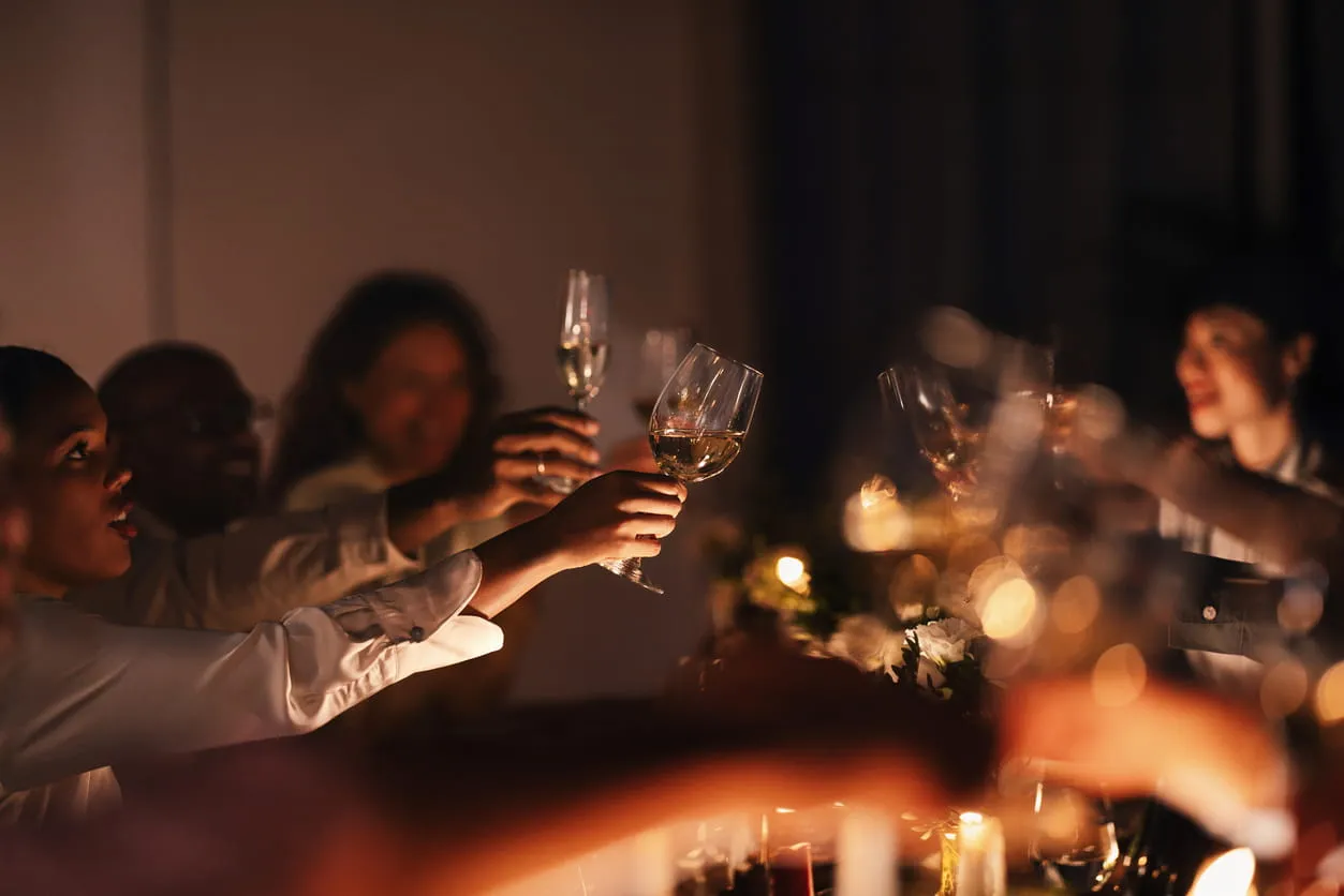 Group of people raising wine glasses in a toast at a dimly lit dinner gathering.