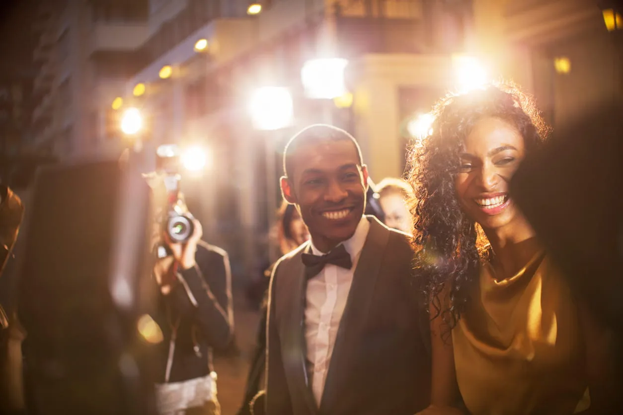Smiling man in a tuxedo and woman in a gold dress at a flash-lit event with photographers in the background.