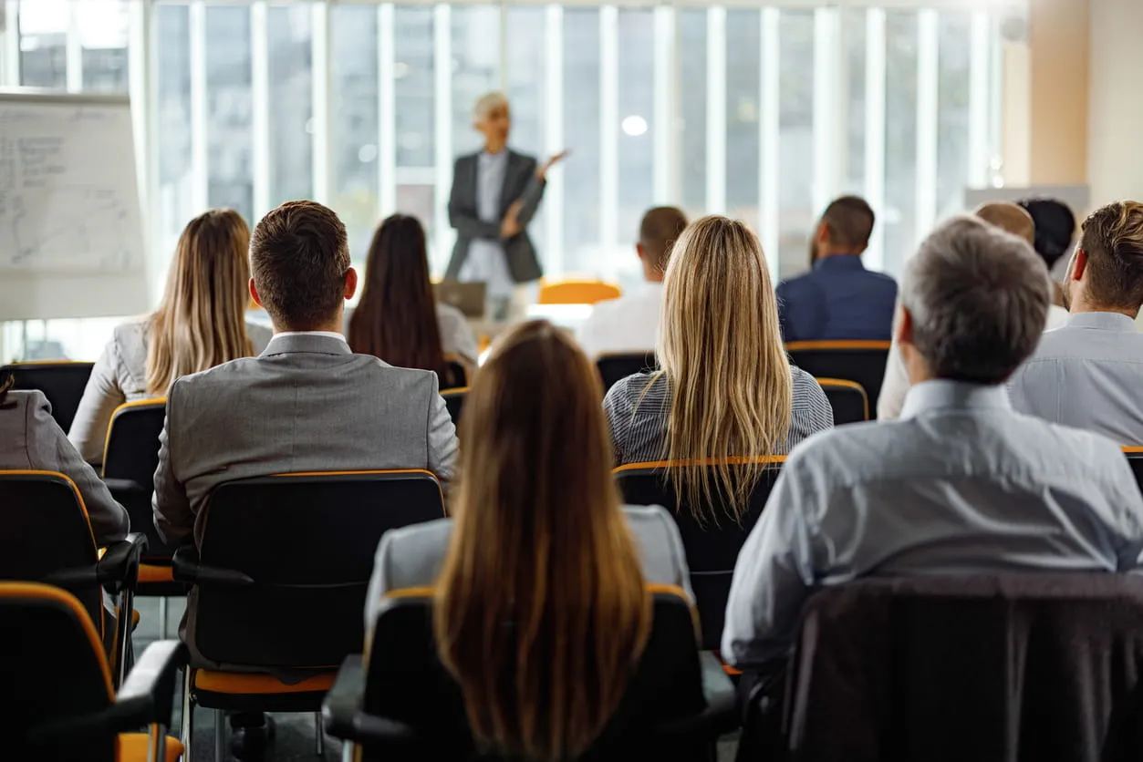 Group of business professionals seated facing a presenter giving a talk in a bright conference room.