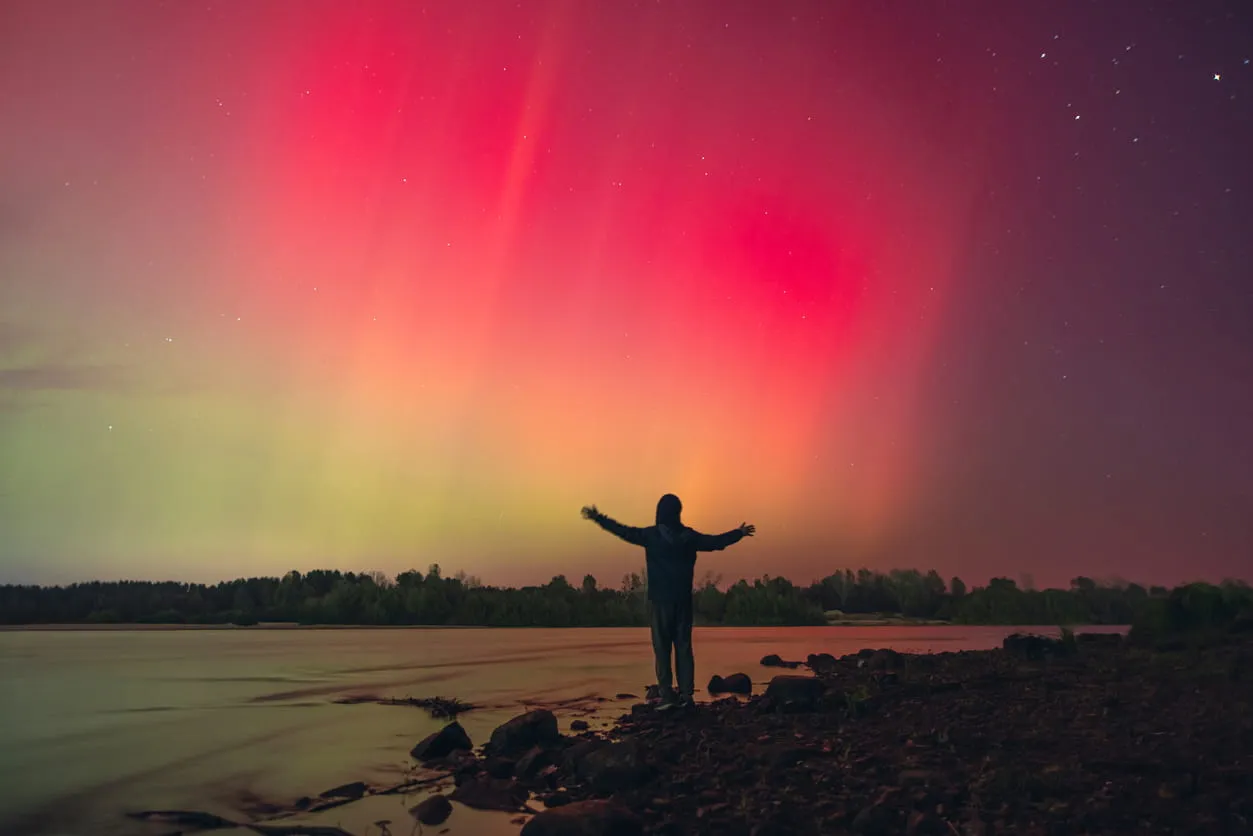Person standing on rocky shore with arms outstretched under a vibrant red and green aurora sky.