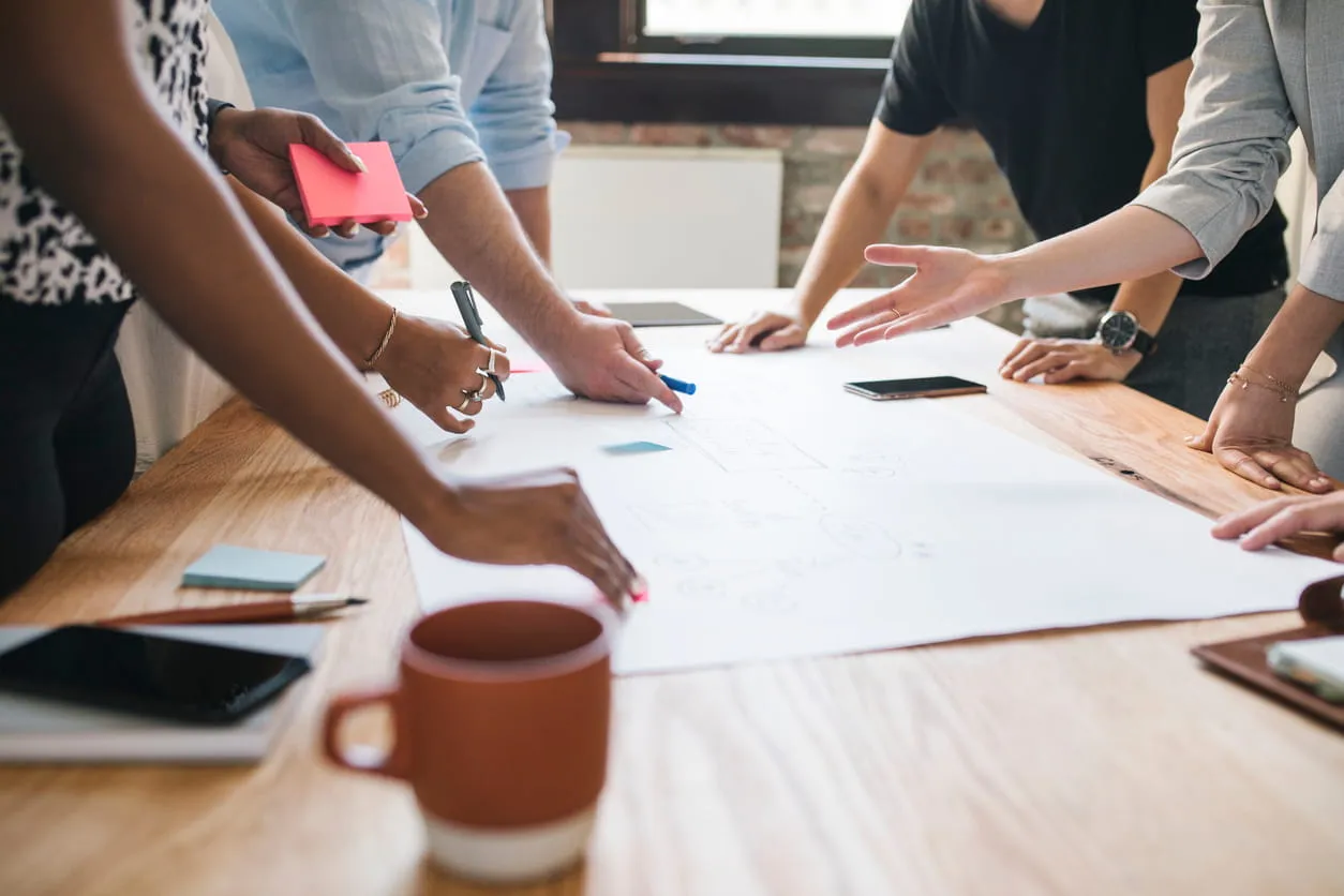People collaborating over large paper on a wooden table with sticky notes and a coffee mug.