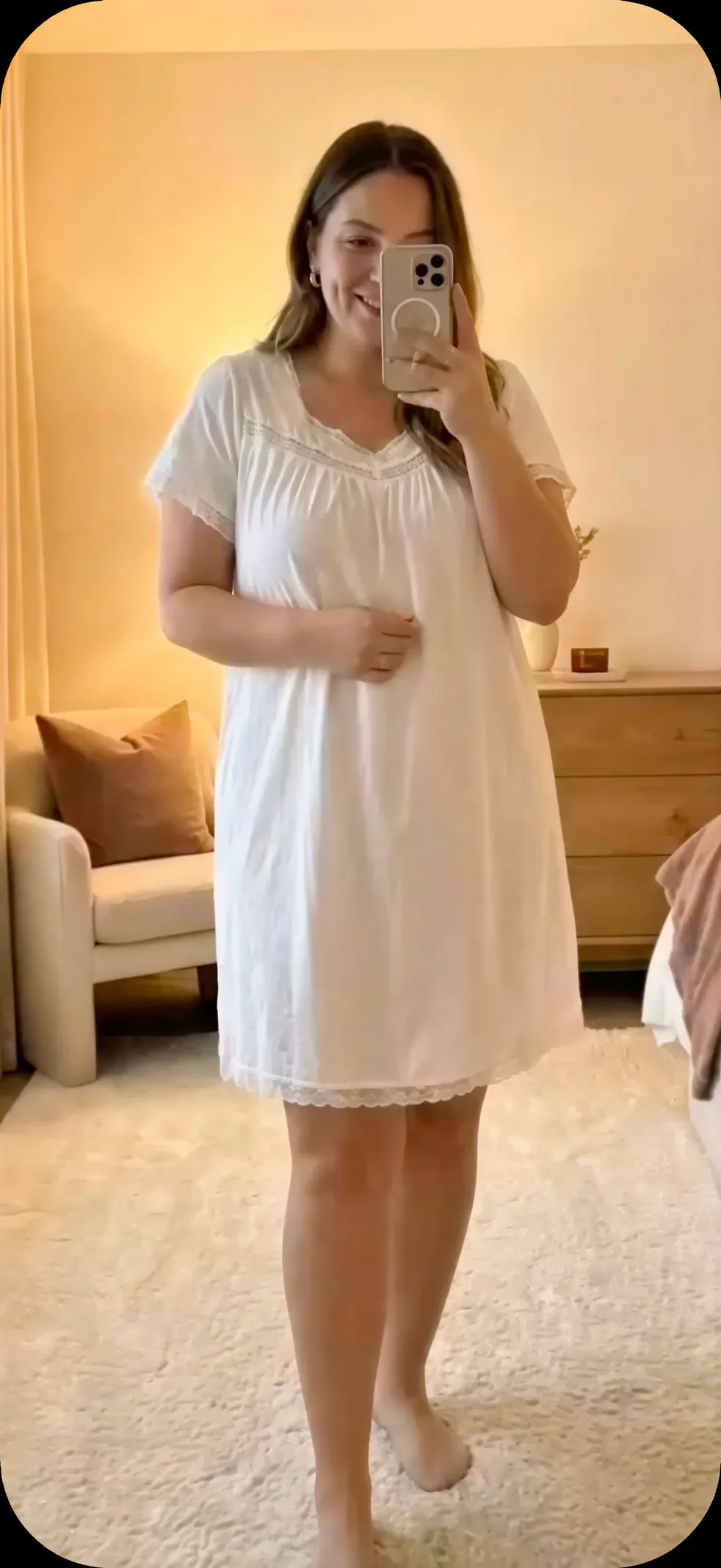 Woman smiling and taking a mirror selfie wearing a white short-sleeve nightdress in a cozy bedroom.