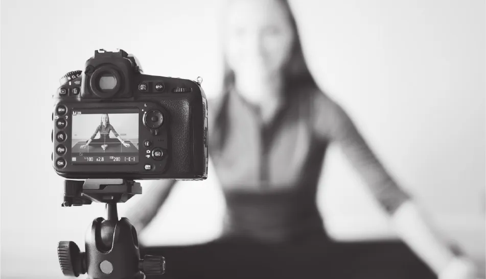 Digital camera on tripod recording a woman sitting cross-legged and smiling in the background.