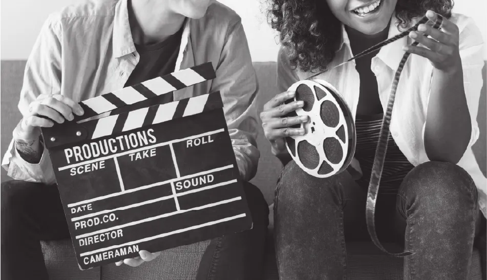 Two people sitting on a couch, one holding a film clapboard and the other holding a film reel with a strip of film.