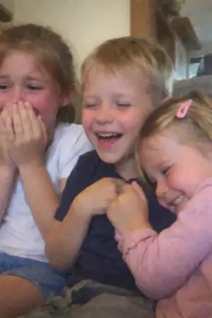 Three young children sitting closely, smiling and laughing together indoors.