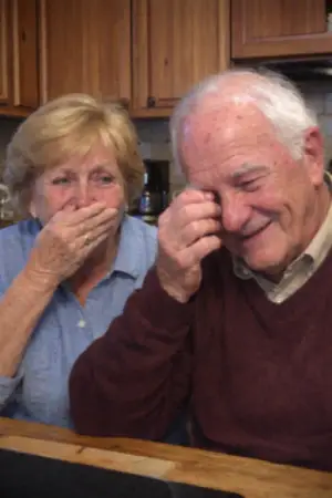 Elderly couple in a kitchen sharing a laugh, the woman covering her mouth and the man wiping his eye with his hand.
