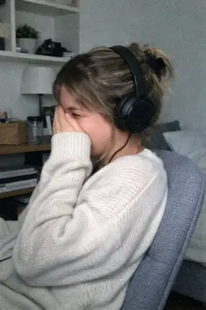 Woman wearing headphones and a cream sweater covering her face while sitting on a gray chair.