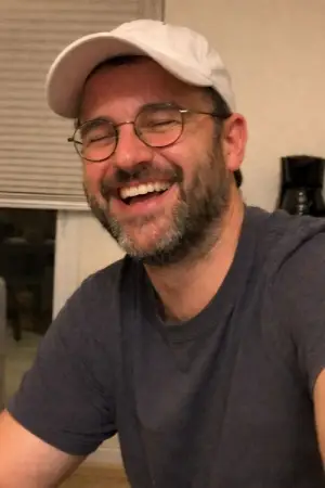 Smiling man with glasses and a white cap, wearing a dark t-shirt indoors.