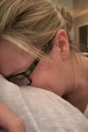 Close-up of a woman with glasses and blonde hair resting her face on a light-colored fabric, appearing distressed.