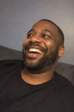 Smiling man with short hair and beard wearing a black shirt sitting against a gray-blue wall.