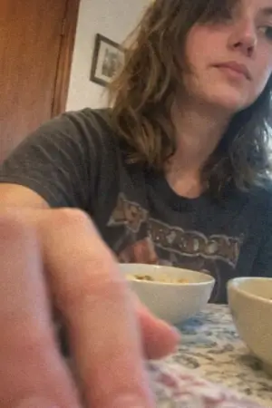 Young person with shoulder-length hair sitting at a table with bowls, hand reaching forward.
