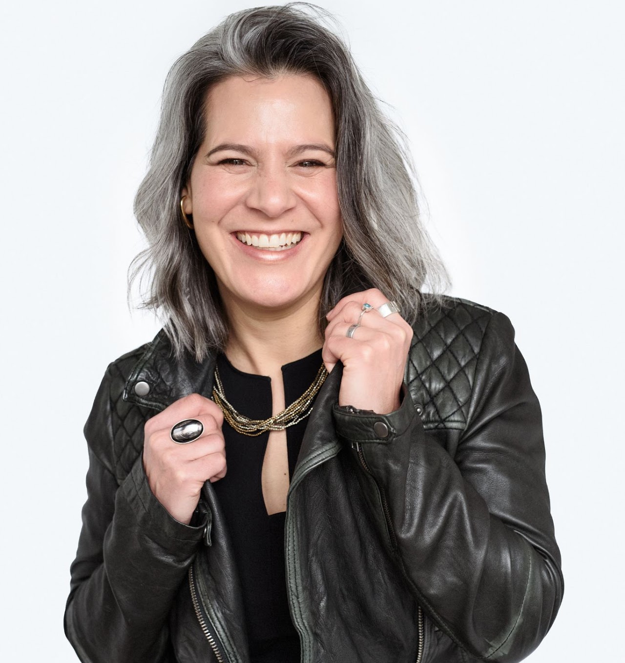 Smiling woman with gray hair wearing a black leather jacket and layered necklace against a white background.