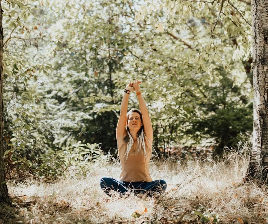 Woman sitting cross-legged on grass in a sunlit forest, stretching arms upward with eyes closed.