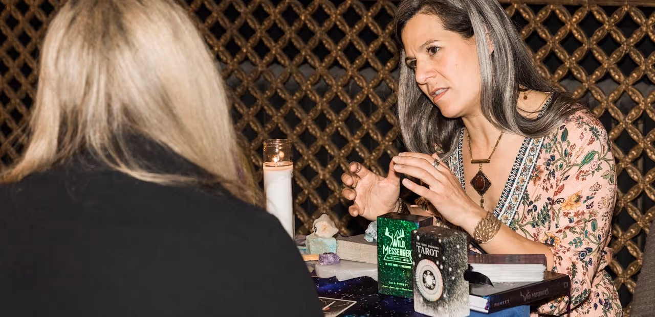 A woman with gray hair and a floral blouse reading tarot cards and crystals to a blonde woman across a table with a lit white candle.