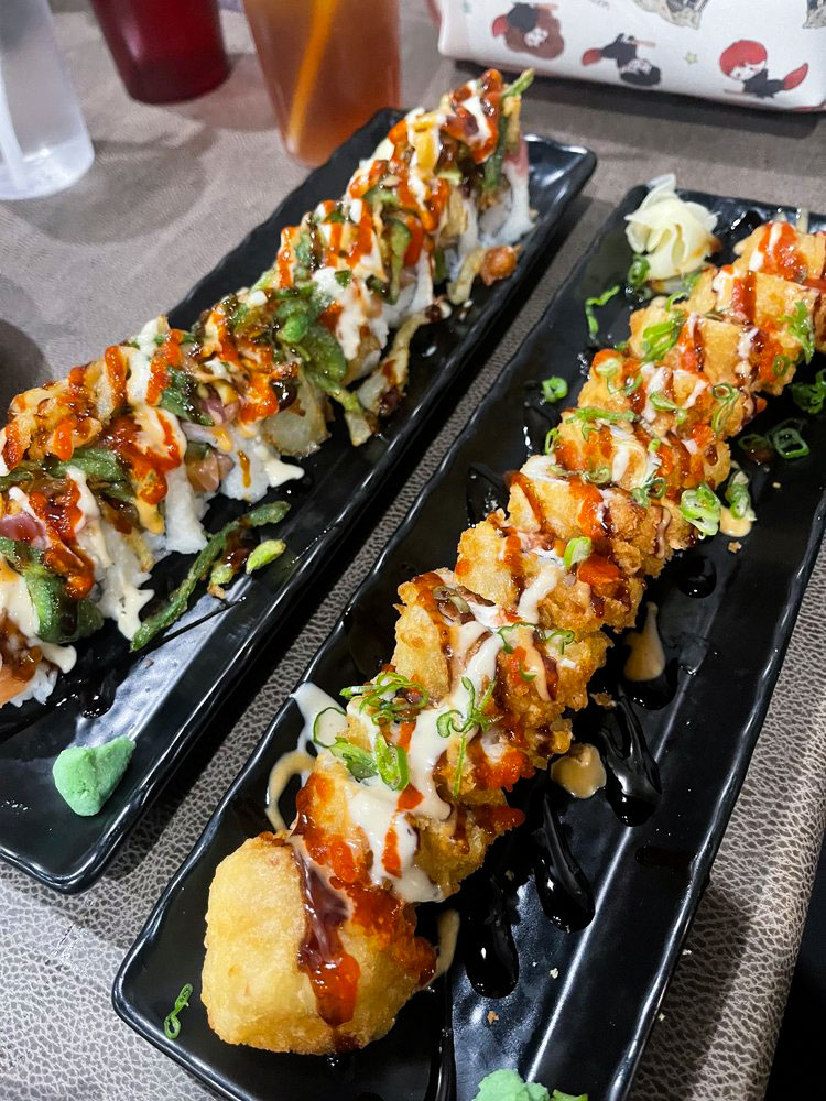 Two sushi rolls on black plates, drizzled with creamy and spicy sauces, garnished with green onions. 