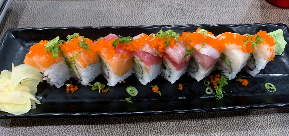A platter of assorted sushi rolls, topped with vibrant orange fish roe and green onion, is served on a black rectangular dish, accompanied by ginger and wasabi.