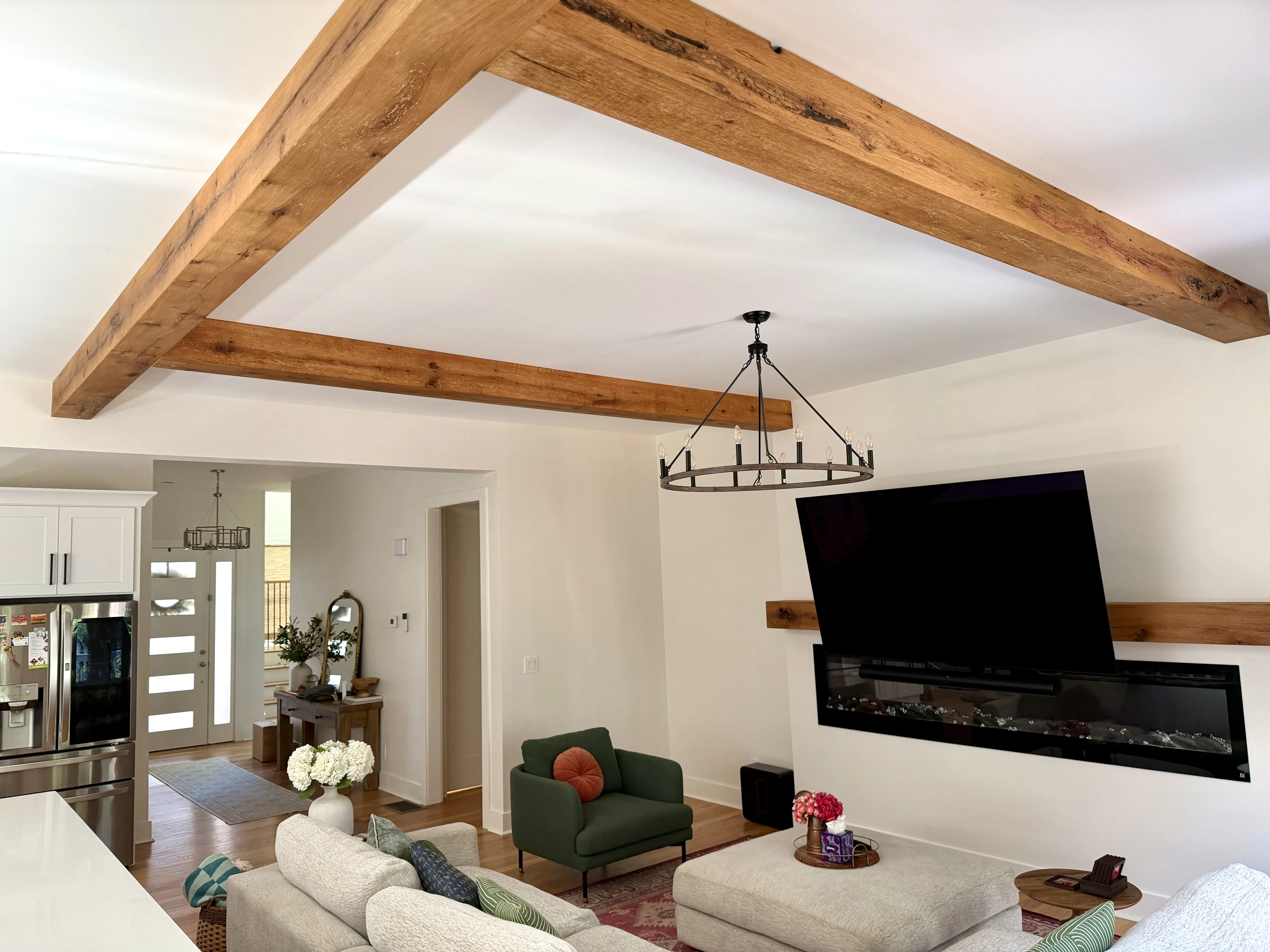 Modern living room with beige sofas, green armchair, a mounted flat-screen TV above a linear fireplace, wooden ceiling beams, and a round chandelier.
