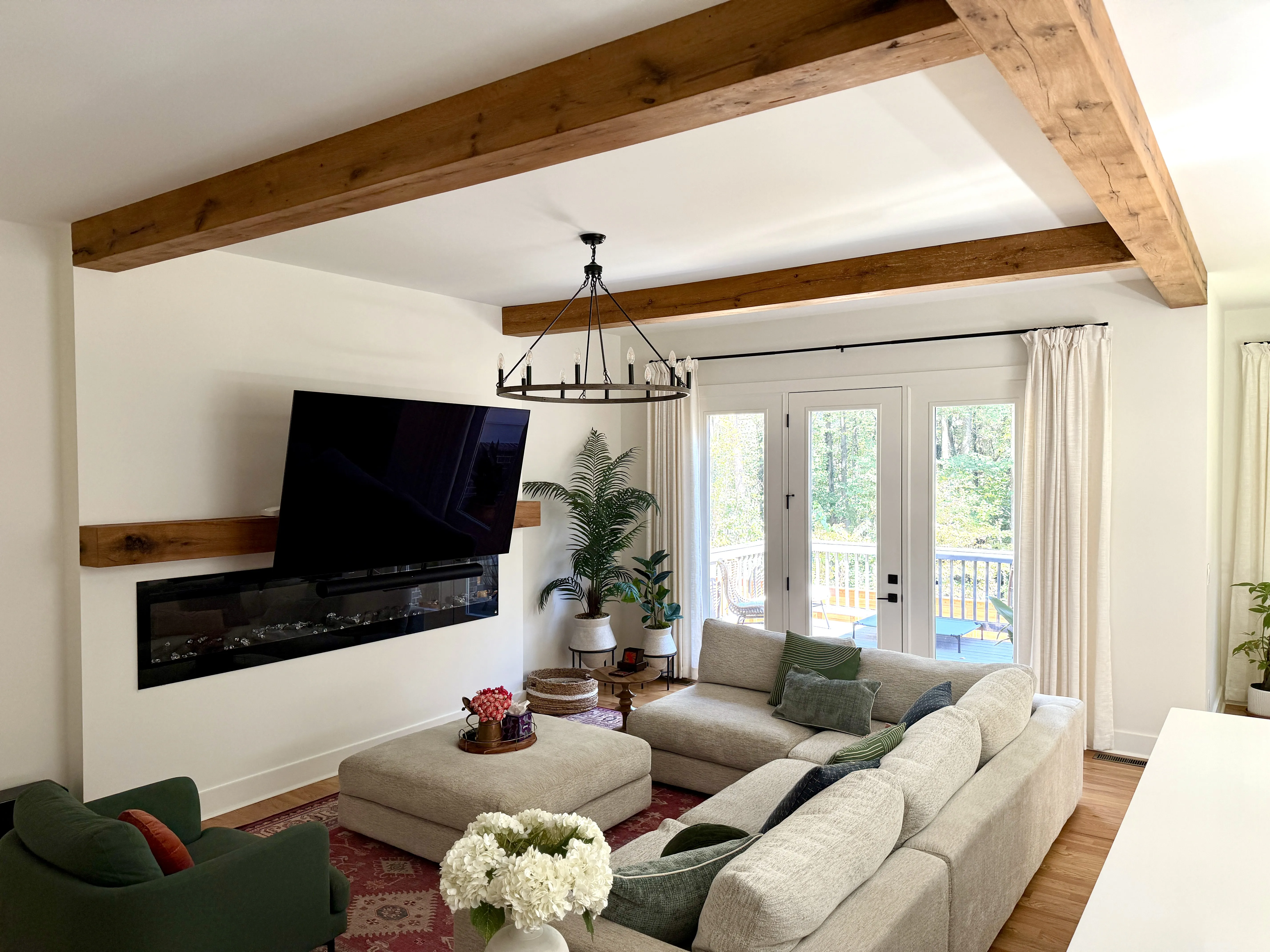 Modern living room with beige sectional sofa, green armchair, large ottoman, wall-mounted TV above electric fireplace, and wood ceiling beams.
