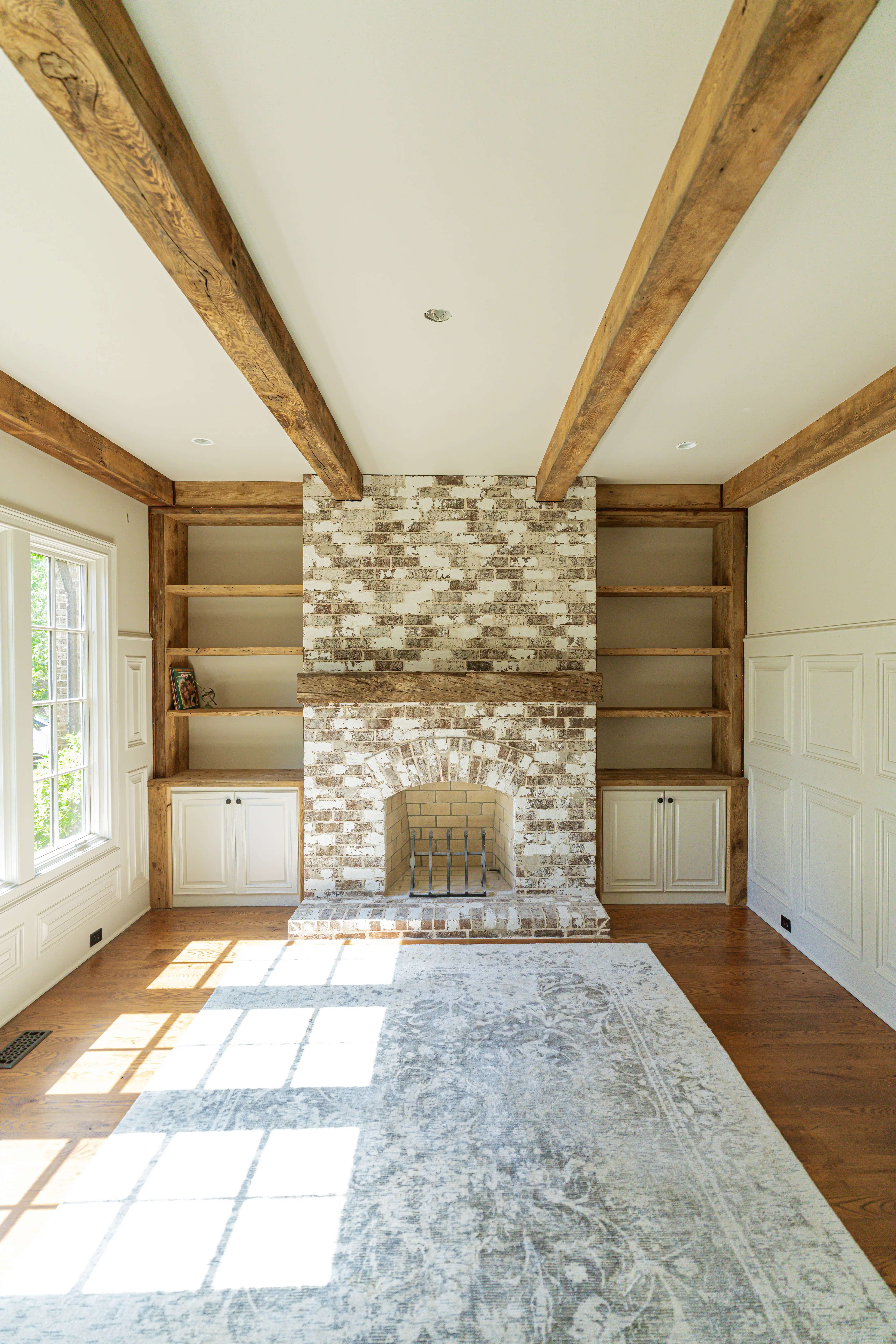 Bright living room with whitewashed brick fireplace, wooden ceiling beams, built-in shelves, and a large patterned rug on hardwood floor.
