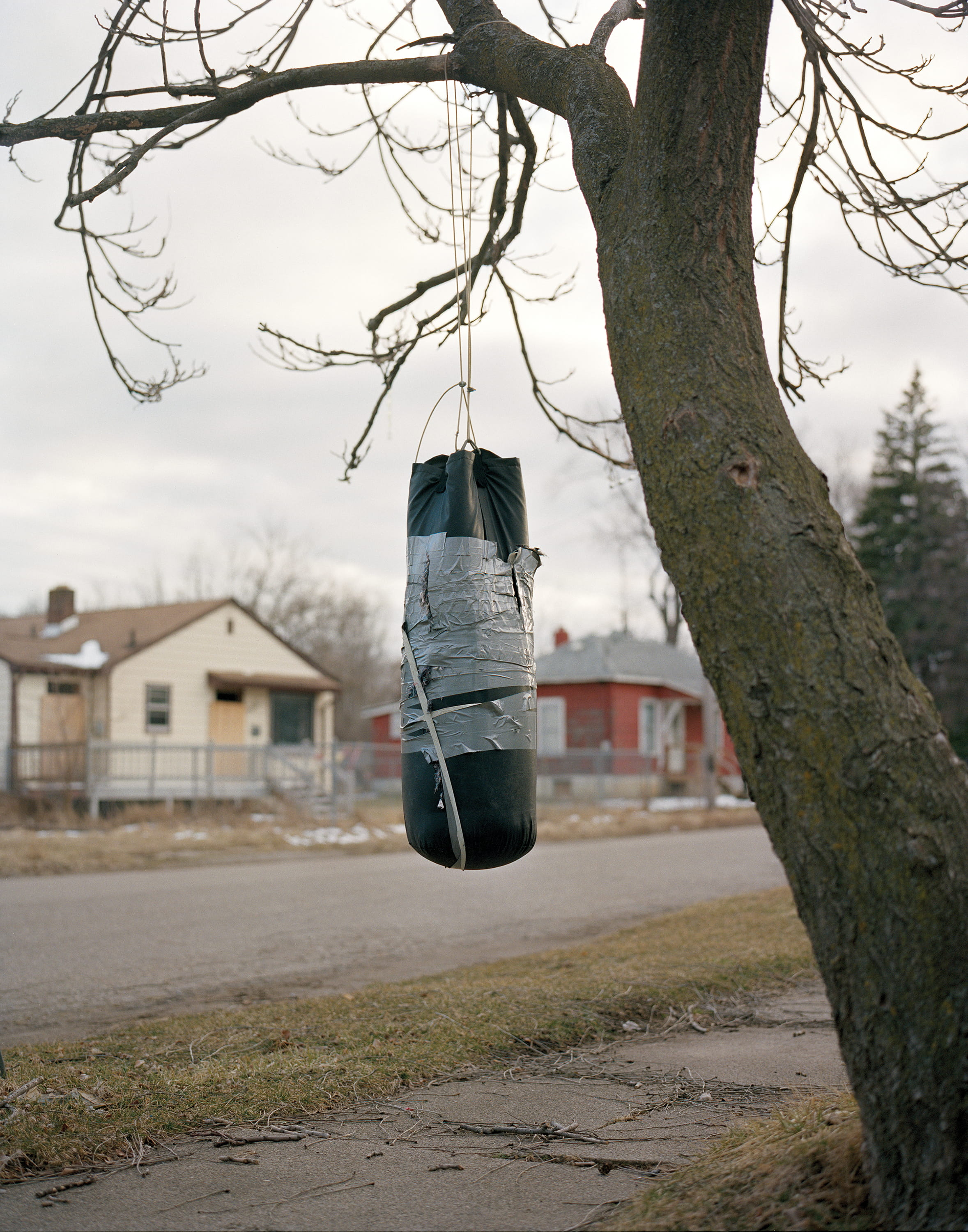 A taped-up heavy bag hangs from a bare tree across from boarded houses