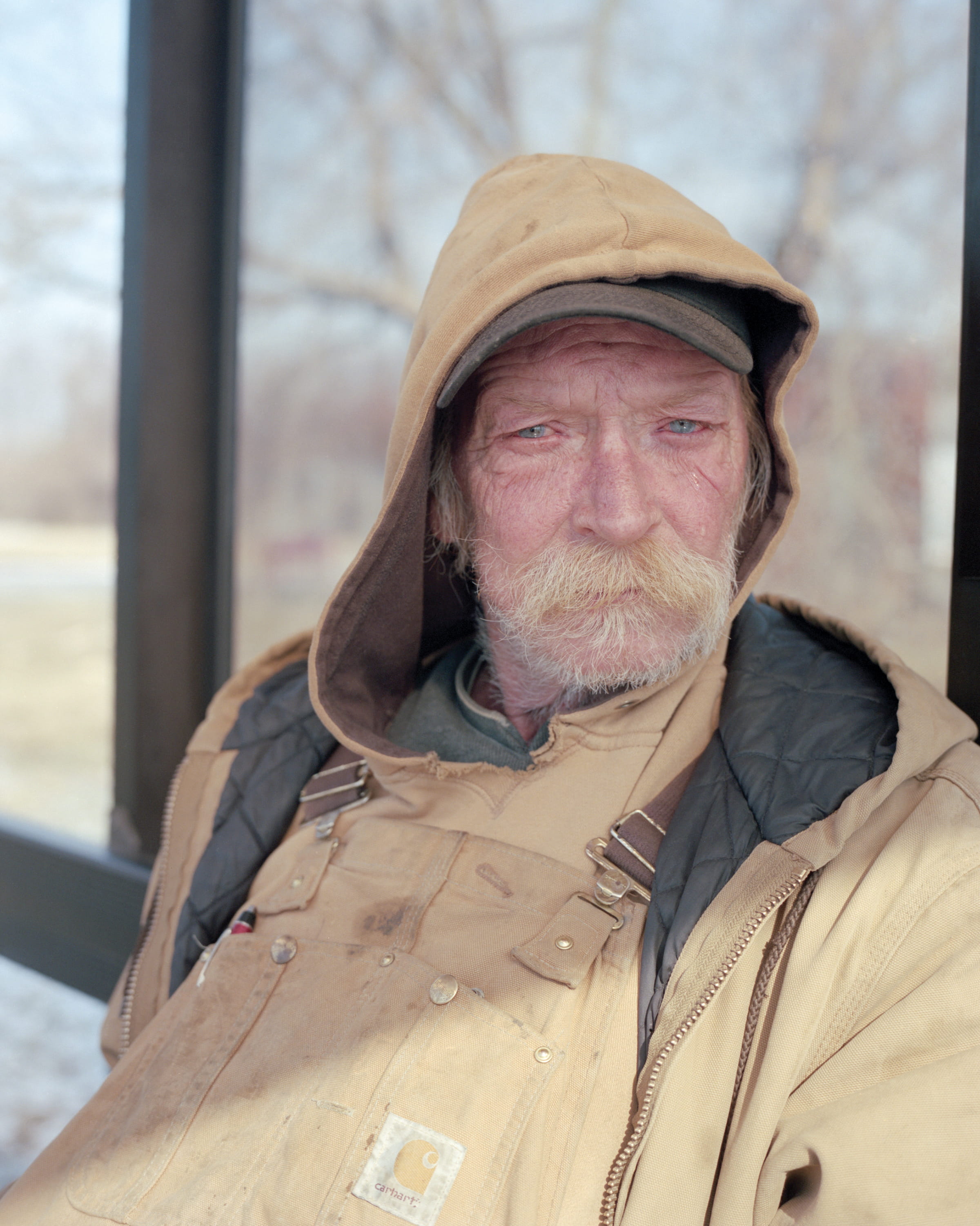 A man in a hooded Carhartt coat waits inside a bus shelter in winter