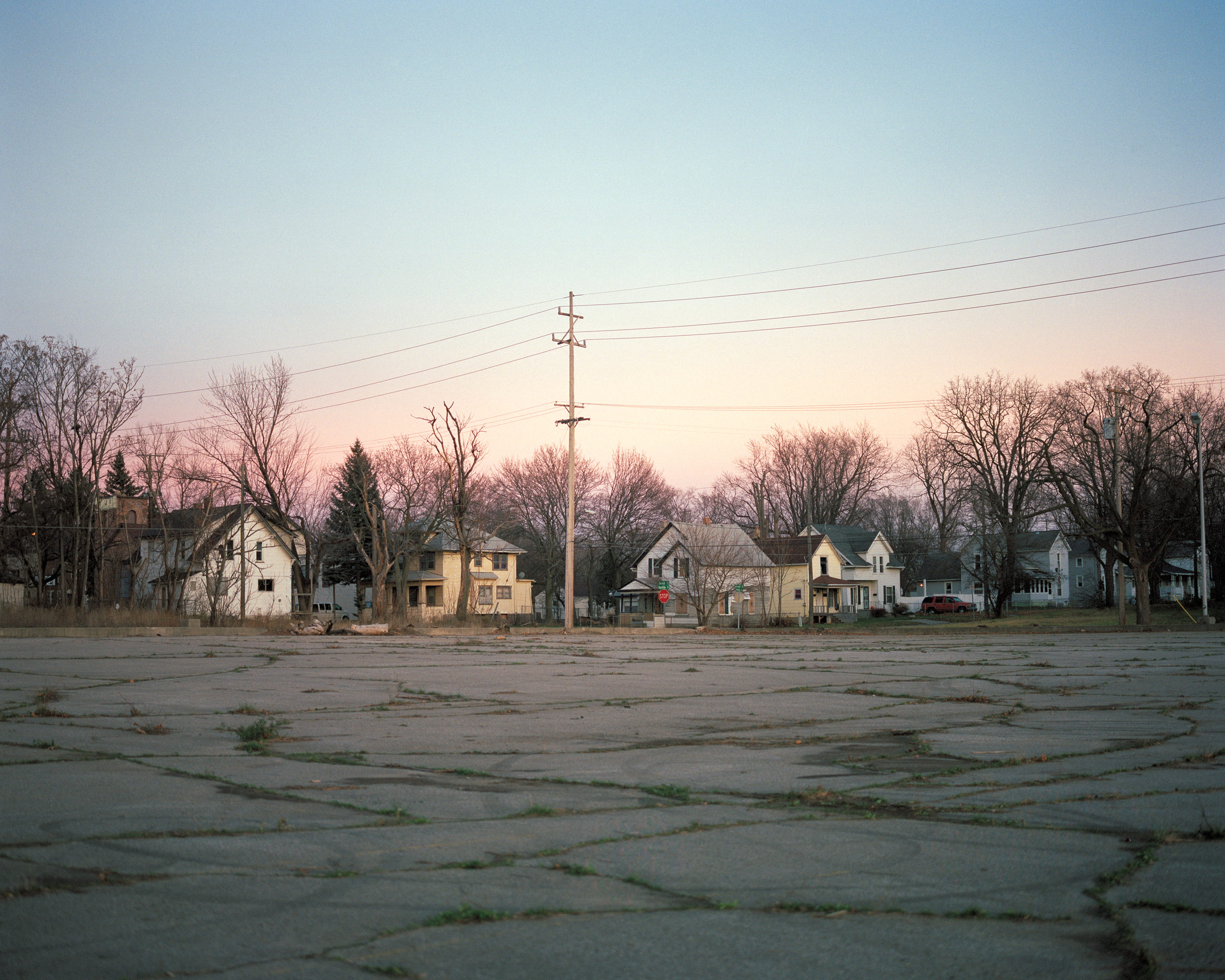 A cracked, empty lot at dusk with houses and power lines beyond