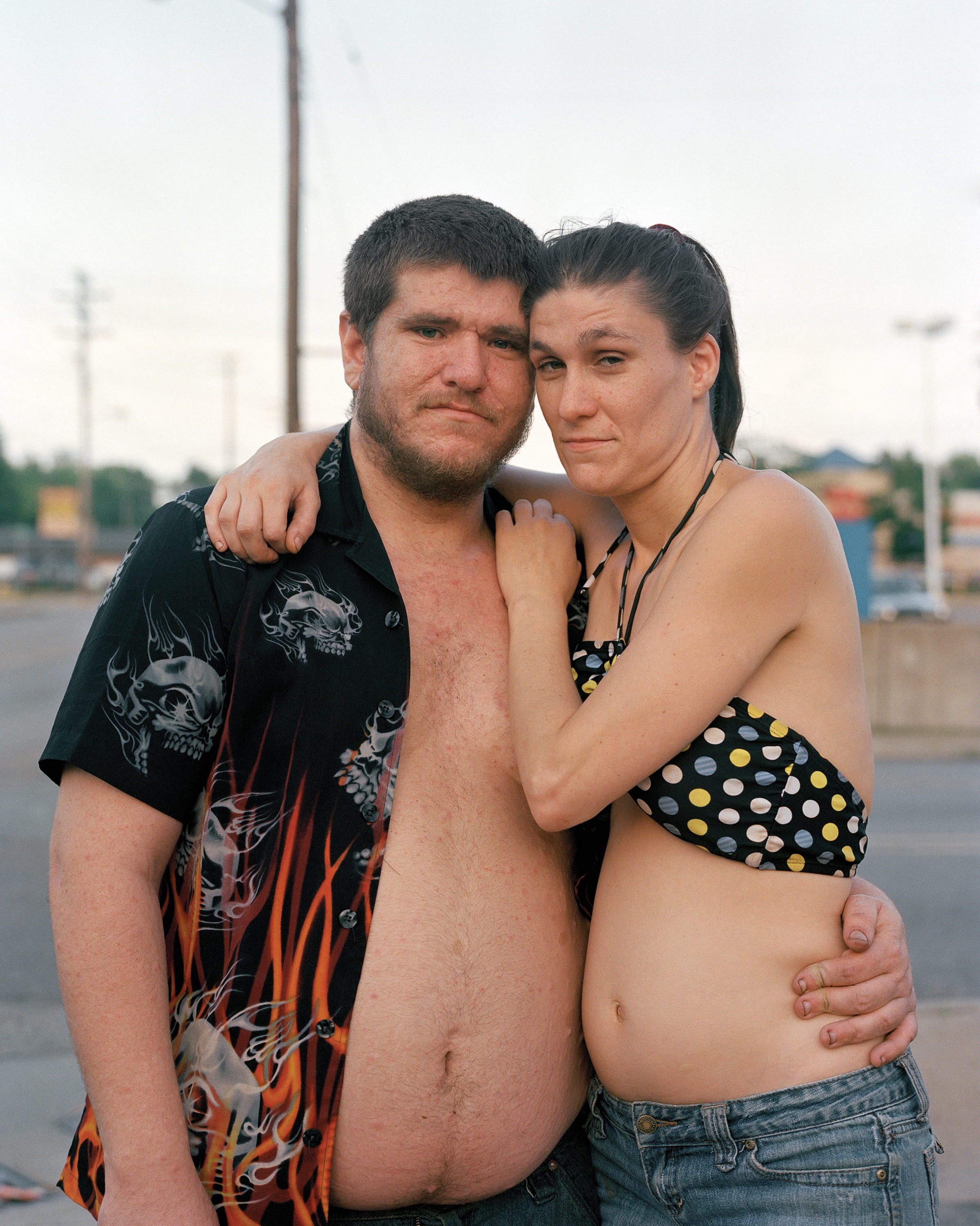 A couple stands together in a parking lot at dusk, arms around each other