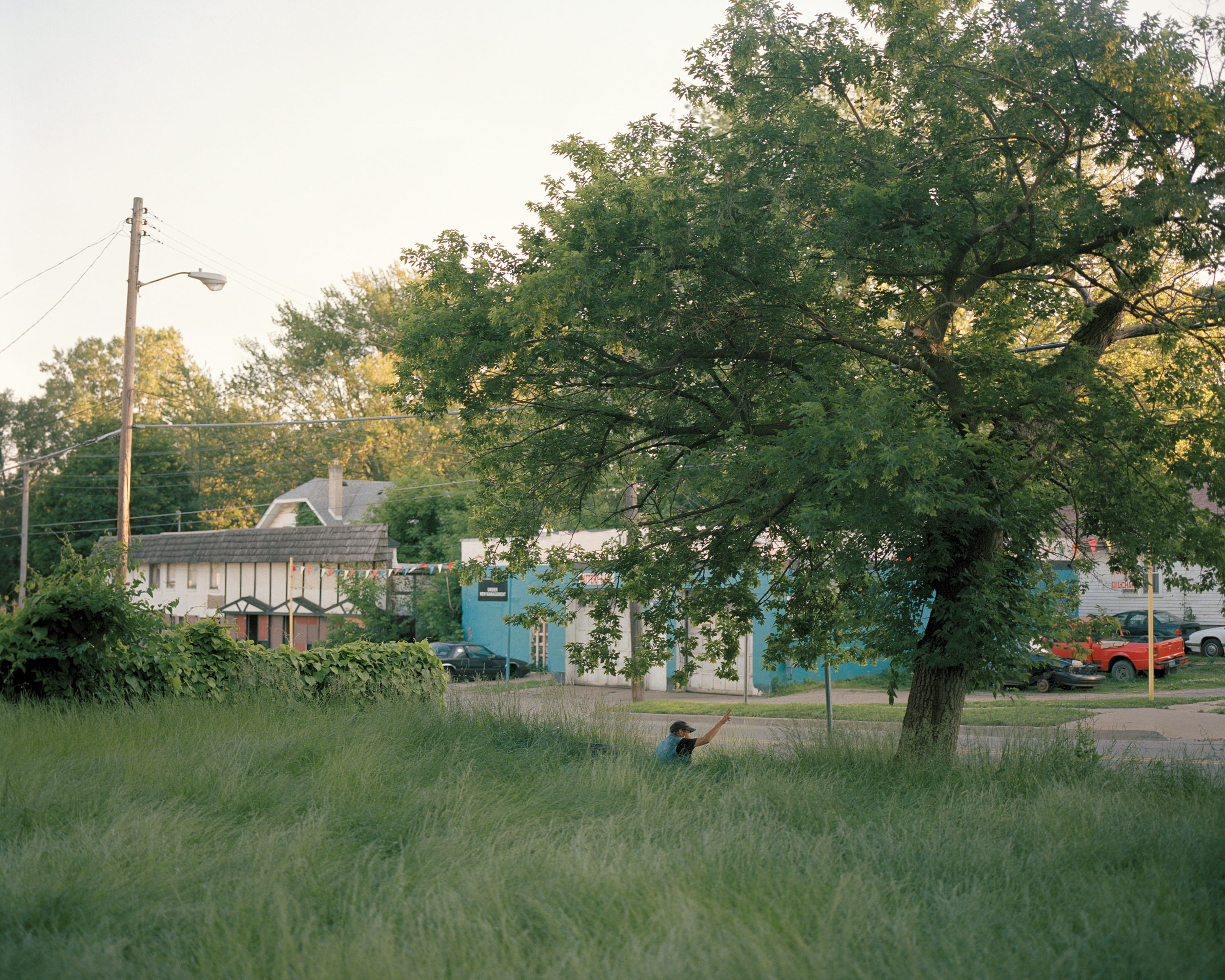 A person raises an arm in tall grass across from a blue storefront, summer evening