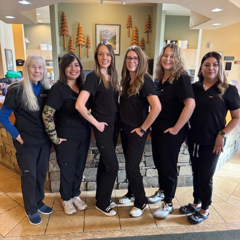 Six women standing inside The Eye Center in Yakima wearing black medical scrubs, smiling for a group photo in a warm-toned room with a stone wall and tree decorations behind them.
