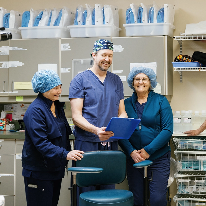 Three medical professionals in scrubs and caps smiling and reviewing a clipboard in a clinical storage room.