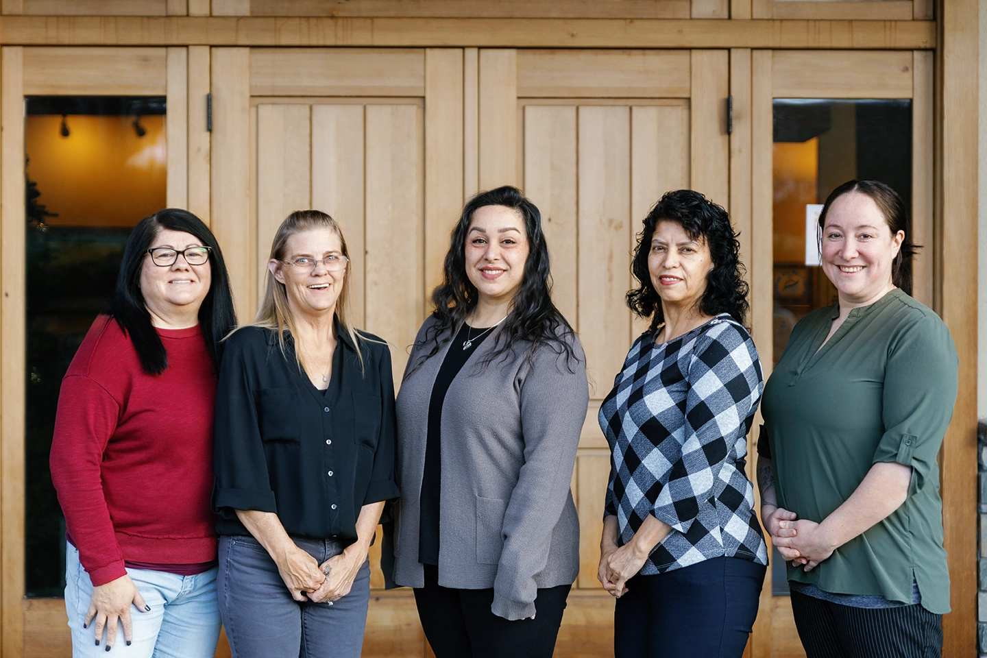 Group of five smiling women standing side by side in front of wooden double doors.