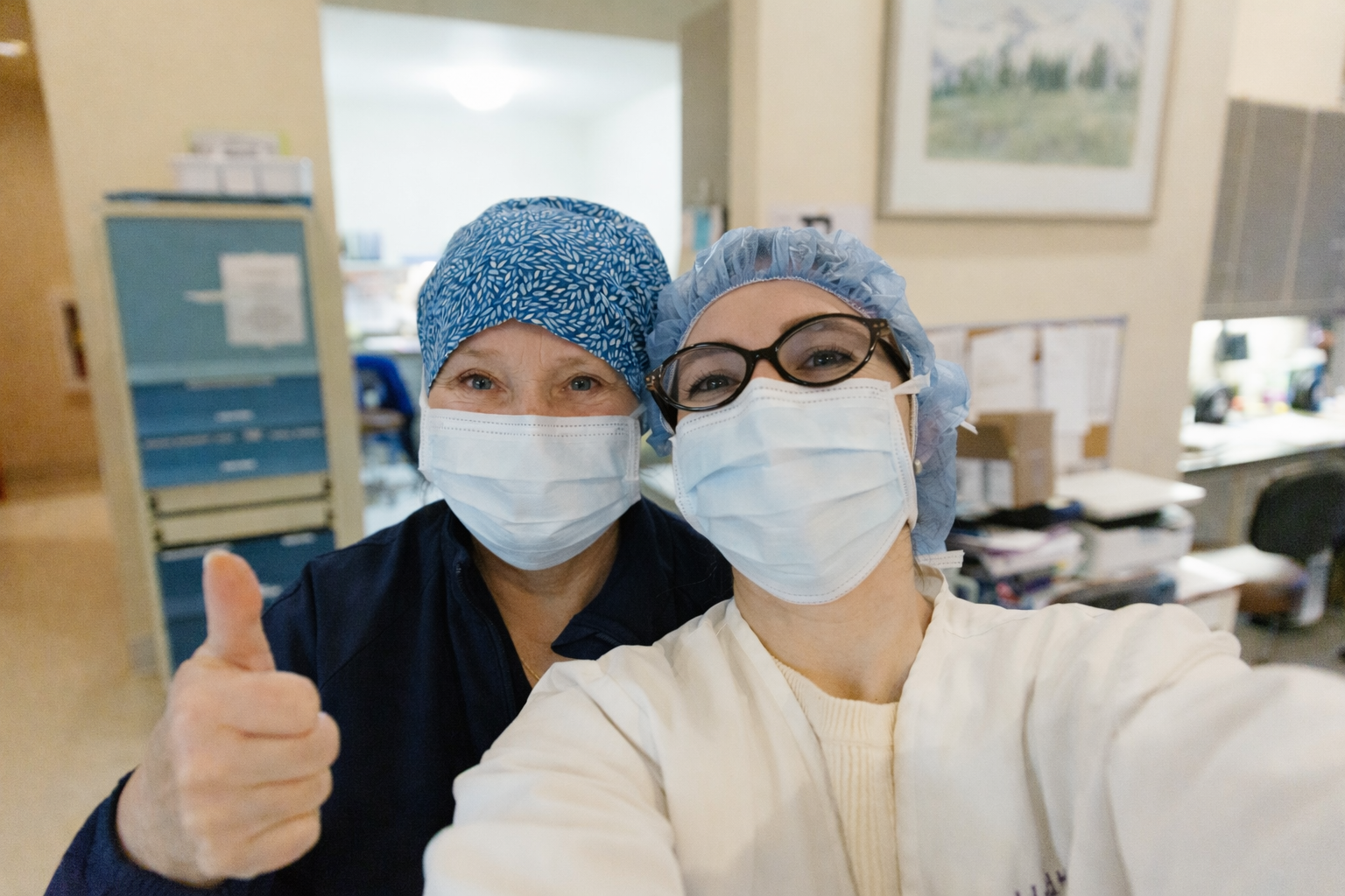 Two healthcare workers wearing surgical masks and caps, one giving a thumbs-up gesture.