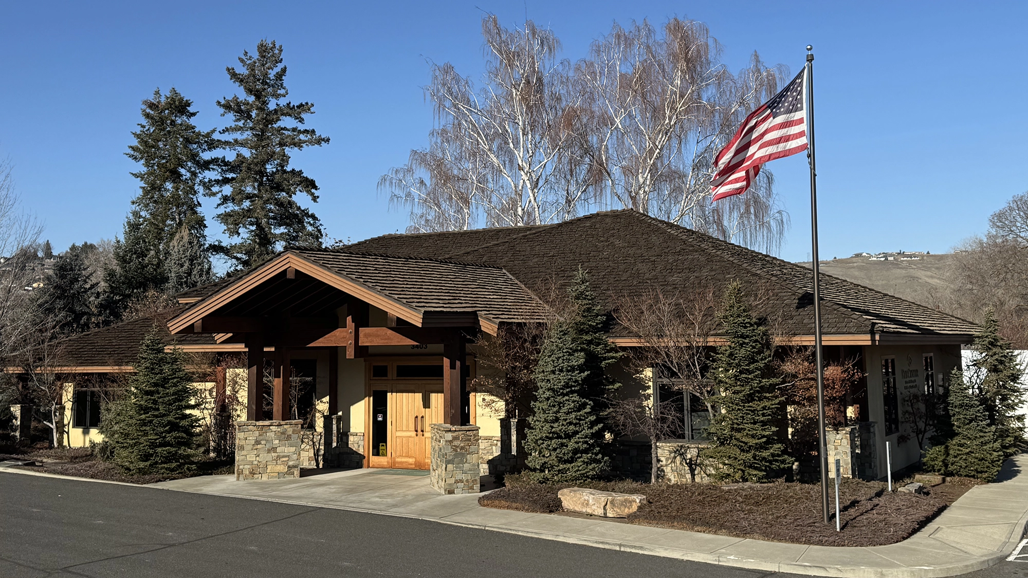 Single-story building with a wooden entrance, stone pillars, evergreen trees, and an American flag on a pole under a clear blue sky. The Eye Center Office in Yakima WA.