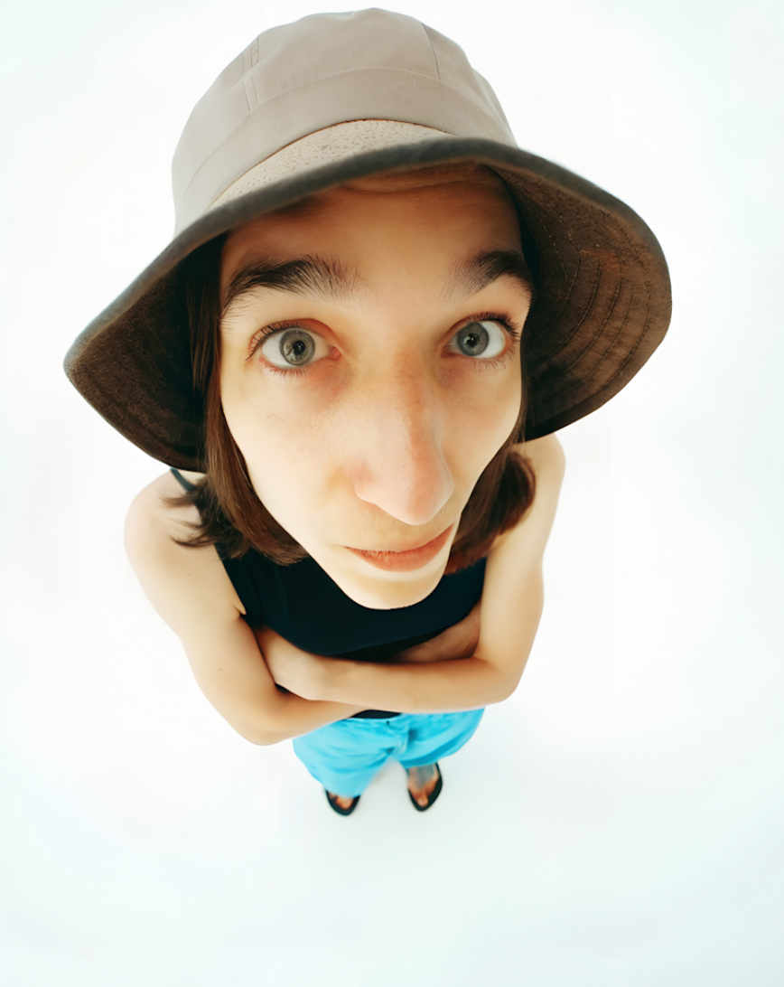 Young person with wide eyes wearing a bucket hat, black tank top, and blue pants standing against a white background.