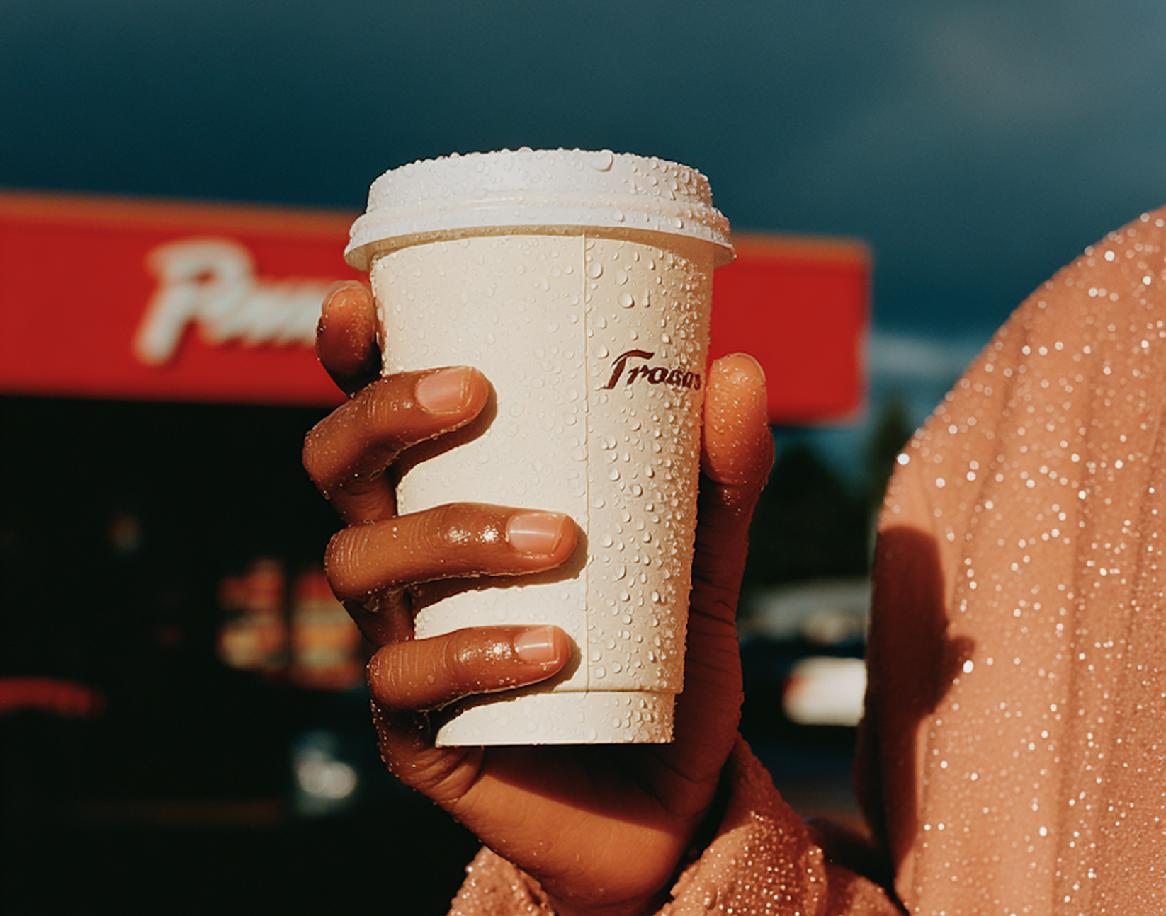 Close-up of a hand holding a white takeaway coffee cup covered in water droplets.