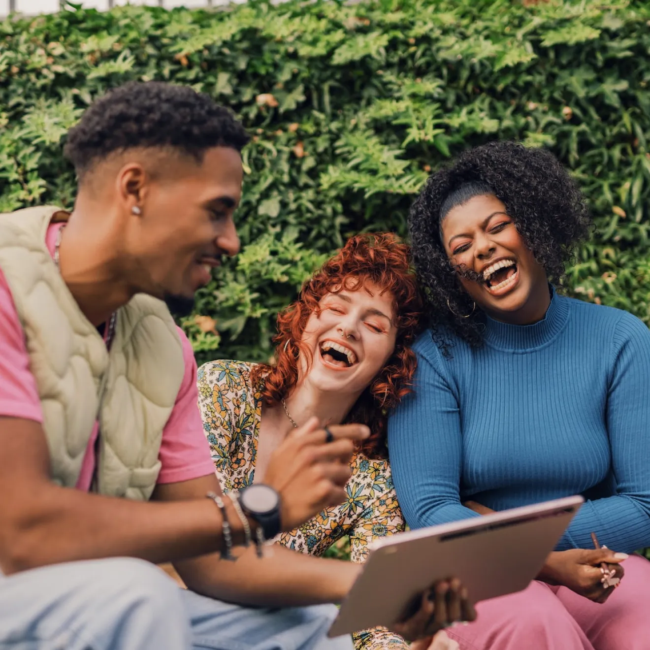 Three friends sitting outdoors laughing and looking at a tablet together in front of green foliage.