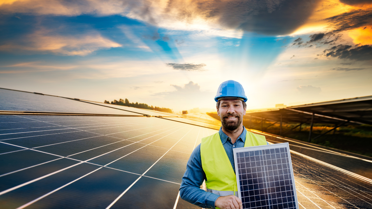 Man in a blue hard hat and yellow safety vest holding a solar panel in a large solar farm at sunset.