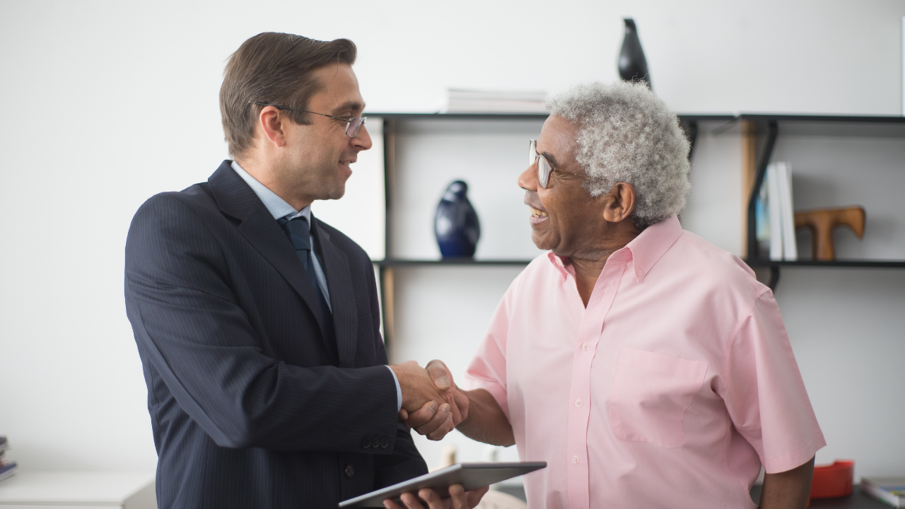 Two men smiling and shaking hands indoors, one holding a tablet, with shelves in the background.
