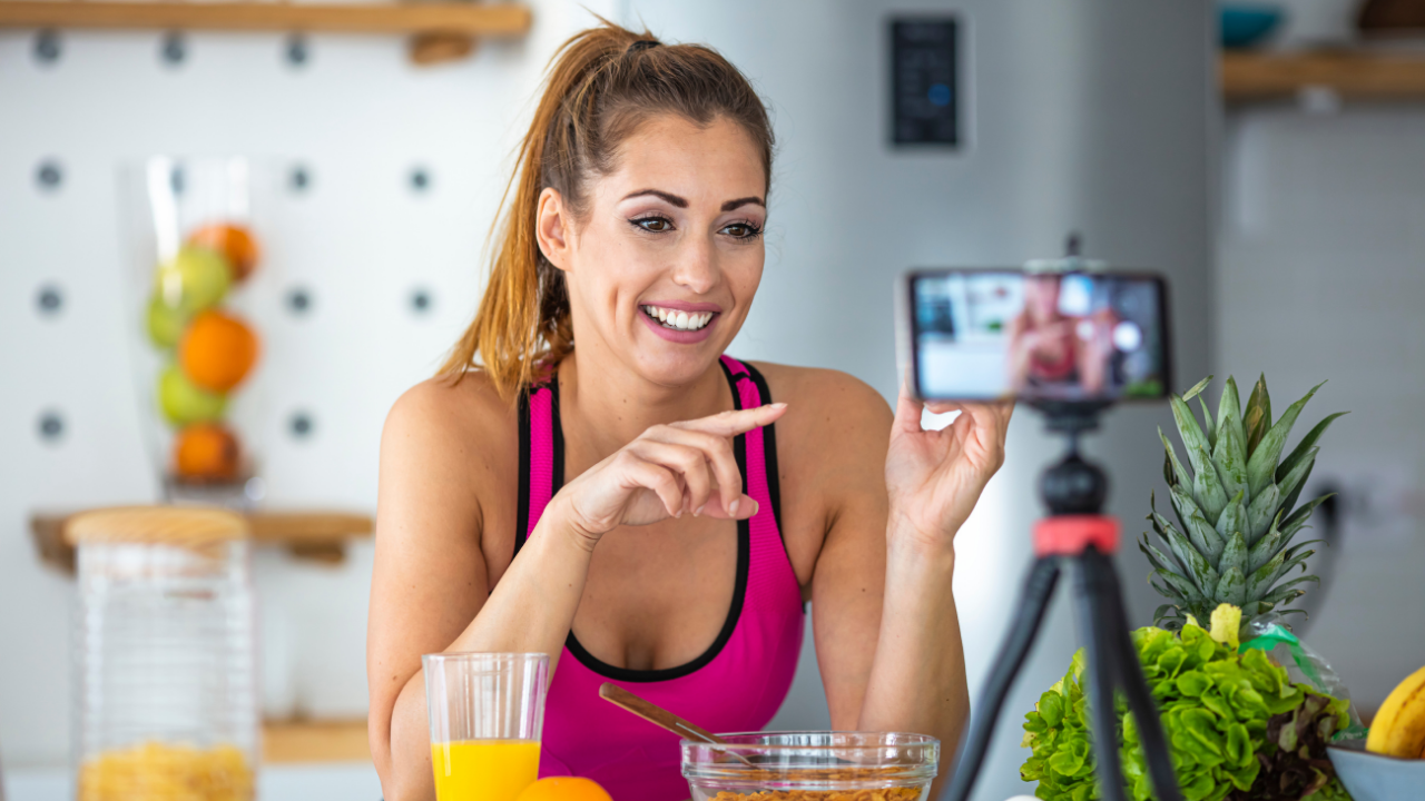 Smiling woman in pink sportswear recording a video in a kitchen with healthy food like pineapple and orange juice.