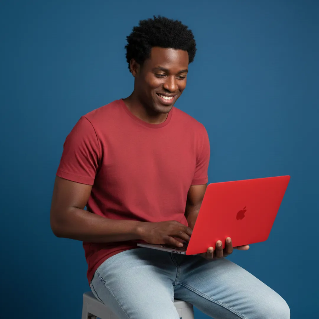 Smiling man in a red shirt sitting on a stool using a red laptop against a blue background.