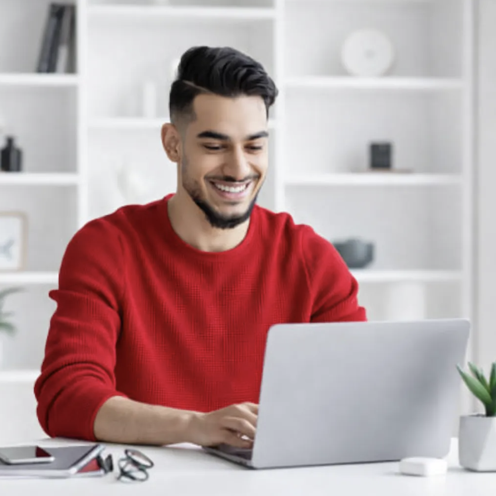 Smiling man in red sweater typing on a laptop at a white desk with glasses, smartphone, and small plant.