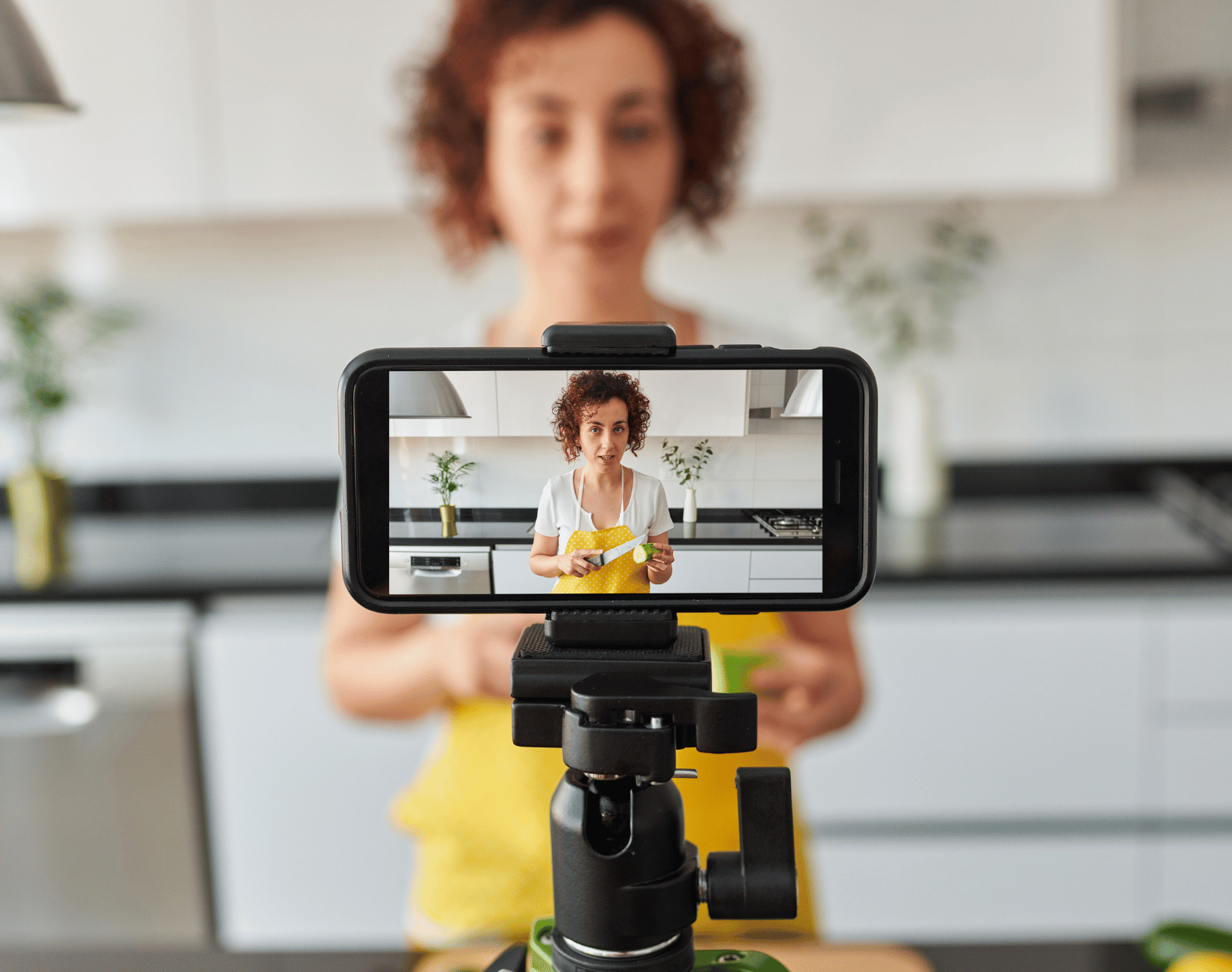 Woman in a yellow apron demonstrating cutting a lime on a smartphone camera set on a tripod in a kitchen.