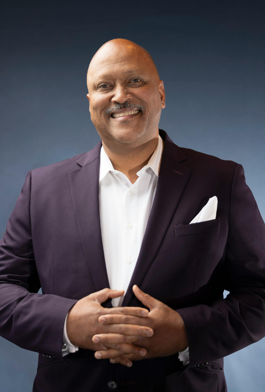 Smiling bald man in a dark suit jacket and white shirt standing with hands clasped in front of a blue gradient background.