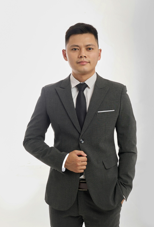 Man wearing a dark gray suit with a black tie and white shirt, standing against a plain white background.