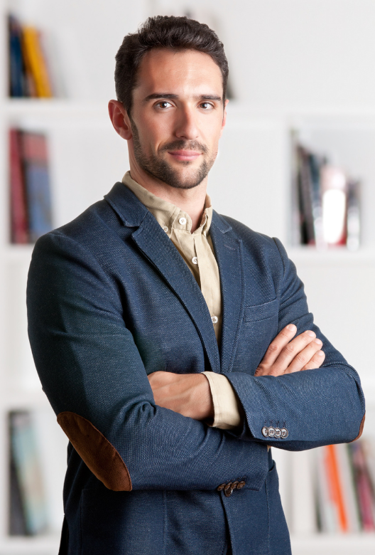 Confident young man with dark hair and beard wearing a navy blazer and beige shirt, standing with arms crossed in front of a white bookshelf.