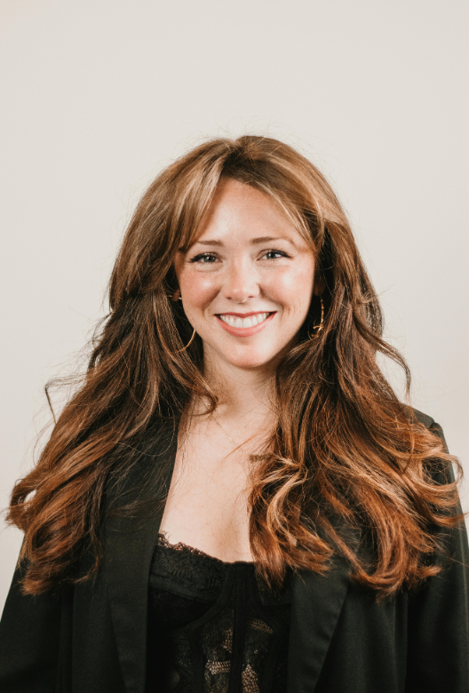 Smiling woman with long wavy brown hair wearing gold hoop earrings, a black lace top, and a black blazer.