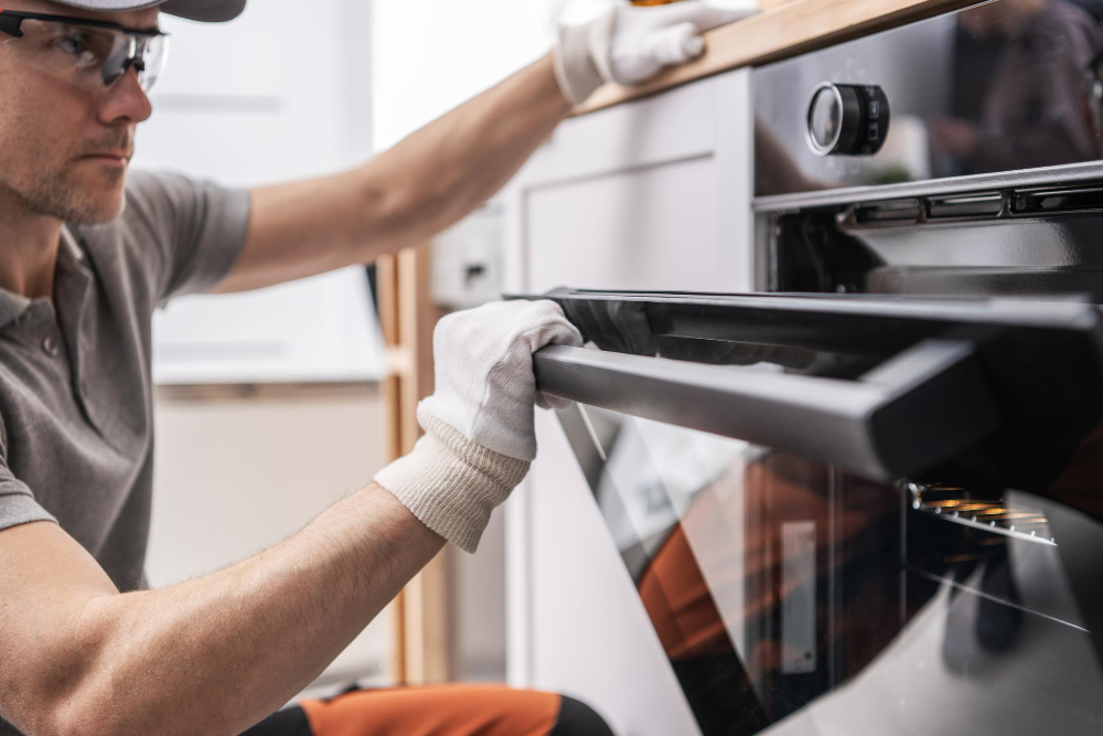 Technician in plaid shirt and overalls inspecting the back of a refrigerator with a red screwdriver in a modern kitchen.