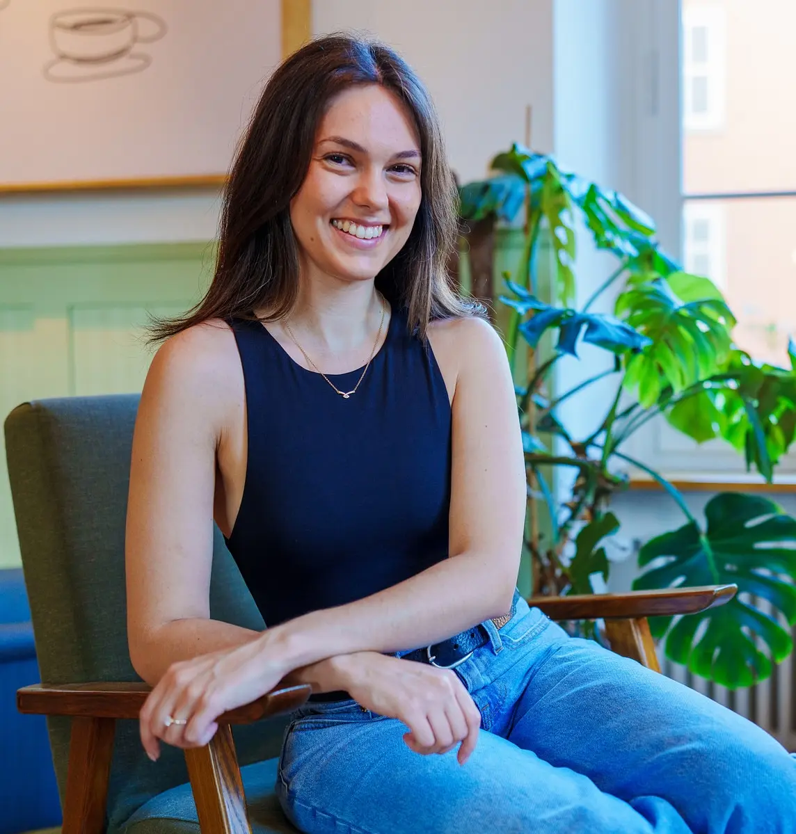 Larissa Sequeira sitzt lächelnd in einem Sessel, im Hintergrund eine Monstera am Fenster