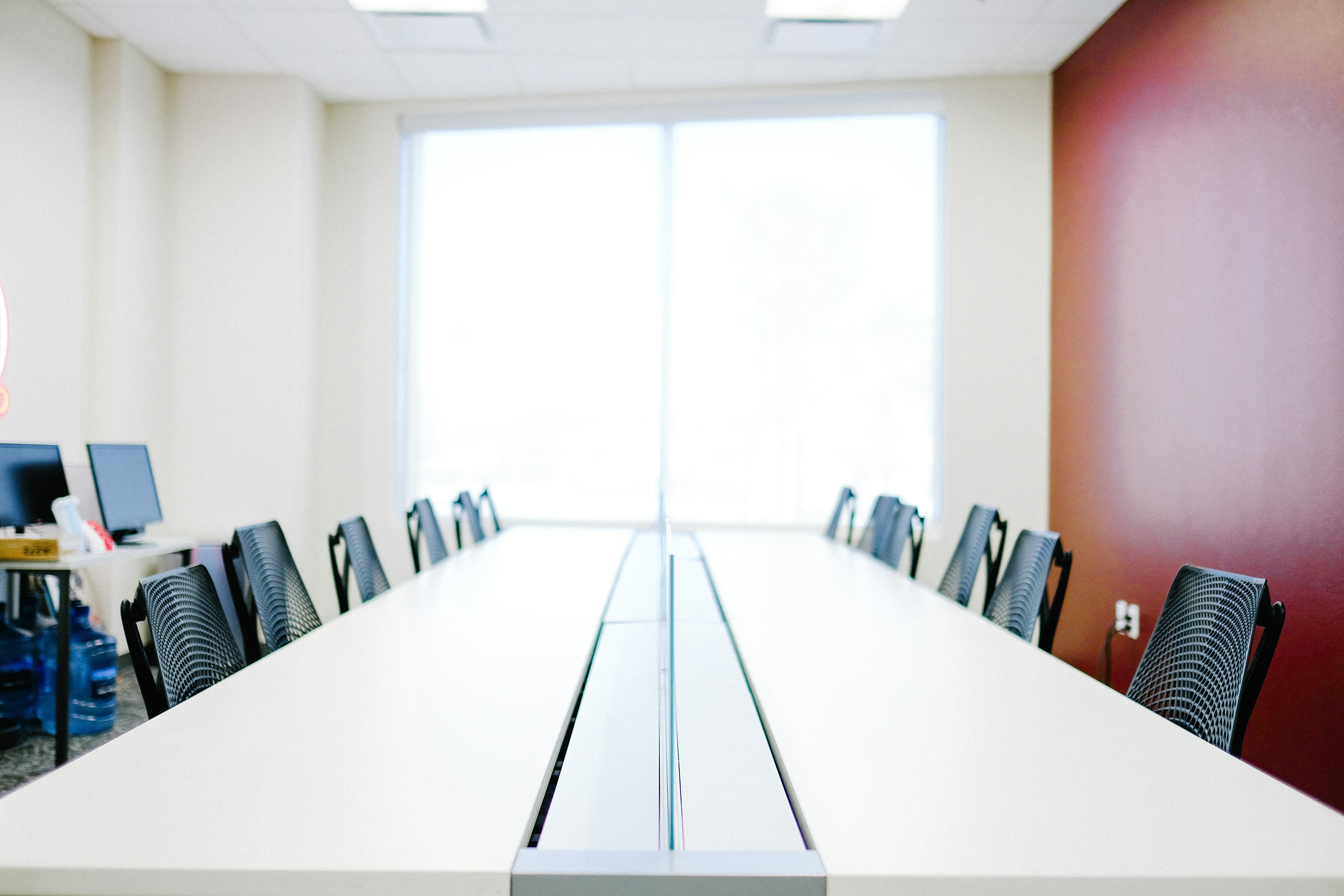 Empty modern conference room with long white tables, black mesh chairs, and large window.